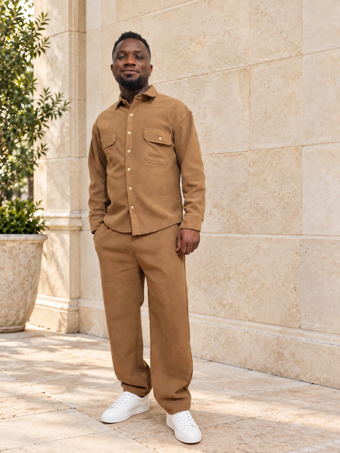 Homme portant l’ensemble URIALEX brun caramel avec des baskets blanches, dans une cour en pierre beige.