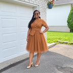 Femme portant la robe URIALEX caramel plissé ceinturée et des escarpins champagne, devant un garage blanc.