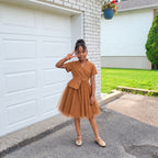 Fille portant la robe URIALEX caramel plissé ceinturée et des ballerines champagne, devant un garage blanc.