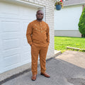 Homme portant l’ensemble tunique et pantalon URIALEX caramel à détails dorés et des mocassins bruns, devant un garage blanc.