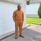 Homme portant l’ensemble tunique et pantalon URIALEX caramel à détails dorés et des mocassins bruns, devant un garage blanc.