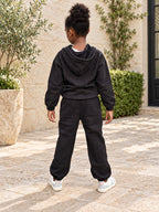 Fille portant l’ensemble URIALEX noir avec haut à capuche et pantalon, vue de dos, dans une cour en pierre beige.
