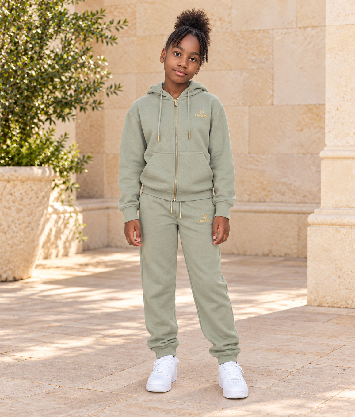 Petite fille portant l’ensemble URIALEX vert sauge, vue de face, dans une cour en pierre beige.